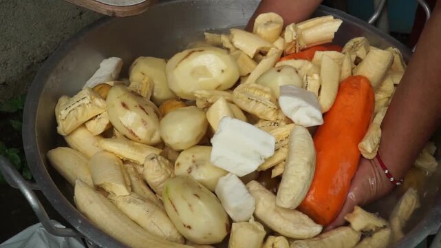 Hands washing chopped traditional Latin American vegetables like green plantains, potatoes, yucca, and carrots in a metal bowl for preparing a Colombian sancocho soup cooking process.