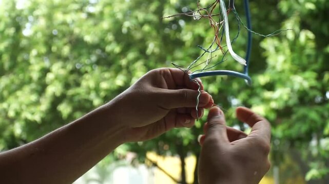 Hands splicing and repairing exposed copper internet wires of a damaged ethernet network cable with a blurry green trees background.