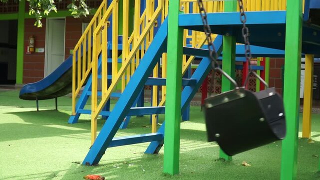 Two empty black rubber swing seats hanging from chains in a colorful kids playground with bright yellow, blue, and green wooden play structures.