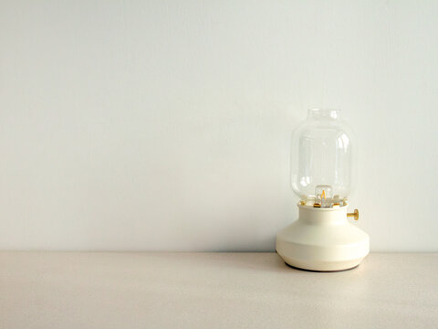 Modern table lantern. Minimalist glass lamp. White wall background. Portable cordless light. Scandinavian interior decor. Clear bulb shade. Ceramic base fixture. Cozy ambient glow. Clean plaster
