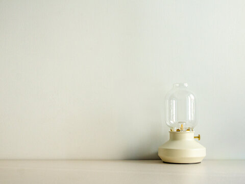 Modern table lantern. Minimalist glass lamp. White wall background. Portable cordless light. Scandinavian interior decor. Clear bulb shade. Ceramic base fixture. Cozy ambient glow. Clean plaster