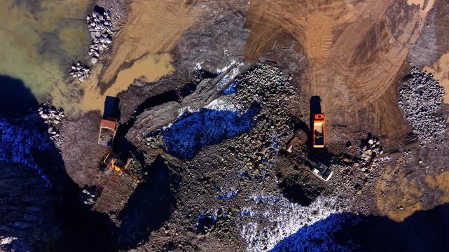 Excavators loading dump trucks. Top down drone shot of two machines beside blue tinted rock and muddy quarry ground.