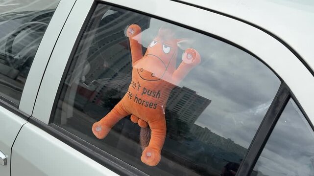 Funny orange toy attached to a car window with suction cups. Quirky travel accessory, humorous decoration, road trip, urban lifestyle, close-up.