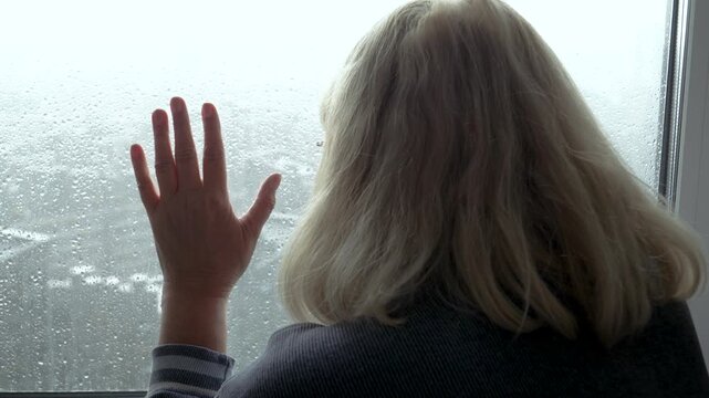 Pensive woman with blonde hair looking out of window with raindrops, her hand resting against cold glass, feeling sad and isolated while watching city during rainstorm. Alone city life in modern world
