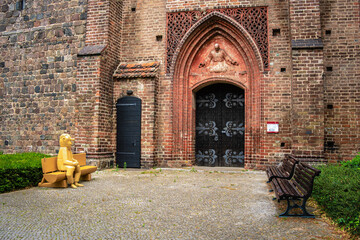 Gothic church portal with an ornate tympanum, flanked by benches and a whimsical wooden figure with a bulbous nose sitting on one of the benches © Margitta