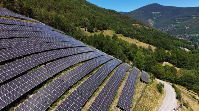 Curved rows of photovoltaic panels follow hillside contours on a forested mountain slope in Andorra, with pine forest, winding road and a valley town visible below