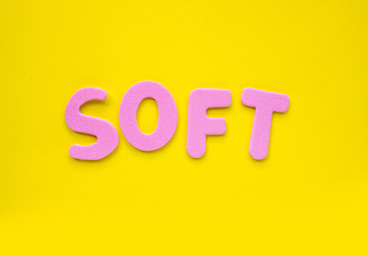 The word 'SOFT' with Pink foam letters on a bright yellow background. Minimal creative flat lay with copy space, representing softness, comfort, typography, branding and modern design concept.