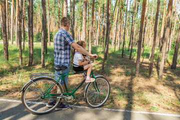 Fototapeta premium Dad rides his daughter in a bicycle seat in forest. A man carries a small child on a bicycle.