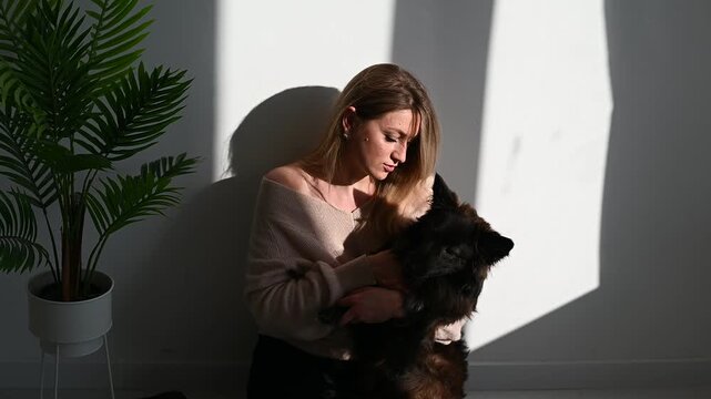 Blonde woman sitting on the floor and playing with a black dog in a bright minimalist room with a potted green plant and natural sunlight.