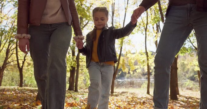 Parents and child walk in autumn park and kick yellow fallen leaves together. Father and mother holding hands of daughter and walking near forest lake, enjoying fall nature and family bonding
