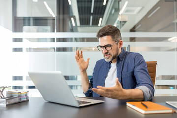 Obraz premium Frustrated man in glasses sitting at a modern office desk, gesturing with his hands while looking at a laptop computer, experiencing stress and technical difficulties
