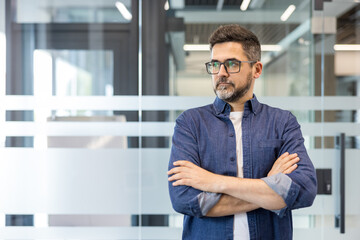 Obraz premium Confident man in glasses with a beard and denim shirt standing with crossed arms looking away, contemplating success within a modern office environment