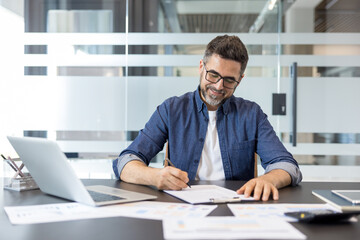 Obraz premium Concentrated businessman in casual attire reading and signing an important contract or document, sitting at a modern office desk with a laptop and paperwork
