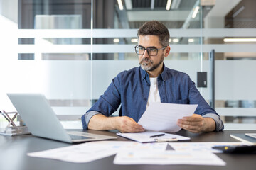 Obraz premium Professional man wearing glasses working with laptop and papers at his desk, analyzing data and preparing reports in a bright corporate environment