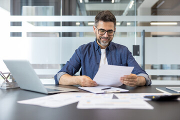 Fototapeta premium Smiling businessman reviewing paperwork and financial reports at his office desk, symbolizing careful planning, corporate success, and meticulous business management