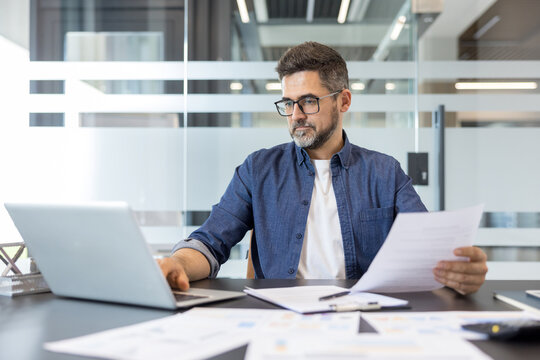 Mature businessman wearing glasses sitting at desk in a modern office, concentrating on his laptop and holding a paper document, representing professionalism, analysis, and career development