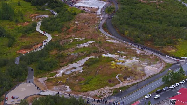 Aerial view of Geysir in Haukadalur, Iceland shows steaming pools, mineral stains, sinuous boardwalks, cars, red roofs, crowds, and rising steam under overcast skies.