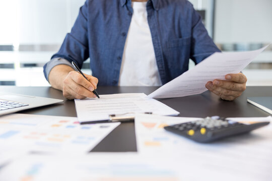 Man in a casual shirt signing a document on a clipboard while holding other paperwork. With a laptop. Calculator. And financial charts scattered on a corporate office desk