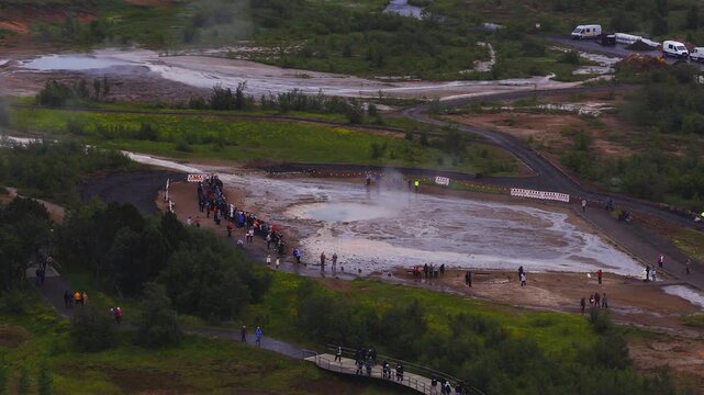 Aerial view of Geysir and Strokkur in Haukadalur, Iceland. Steam rises, visitors queue by boardwalks and platform, vans park, pale deposits meet green tundra in overcast light.