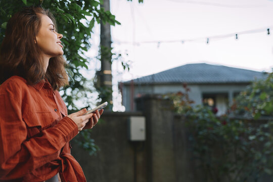 Cheerful woman outdoors, casual lifestyle moment, looking away, soft breeze, warm sunlight, orange sweater, smartphone in hand, relaxed pose among greenery, today