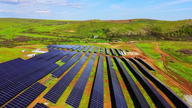 Drone shot of solar farm rows in green valley. Diagonal panel arrays lead toward a distant hillside village surrounded by rolling hills.