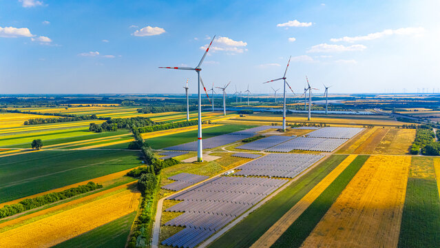 Wind turbines and solar panels in a green field on a sunny day. A large area with numerous wind turbines and solar panels under a clear sky. Fields stretch in different colors.