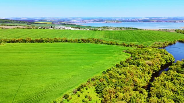 Aerial view of green farmland beside a lake, Bulgaria. Cultivated fields border dense woodland with a river bend and distant industrial site visible.