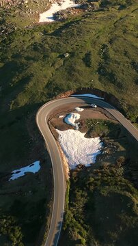Vertical Screen: Beartooth Highway curves through Montana USA alpine landscape with melting snow patches and green tundra as white car navigates hairpin turn at golden hour.