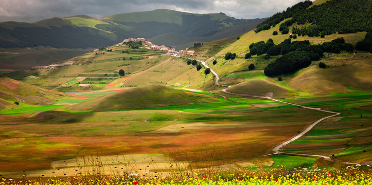 A small village sits atop a rolling green hill in a vast valley. Winding roads traverse the agricultural landscape under dramatic clouds in this scenic mountain area.
