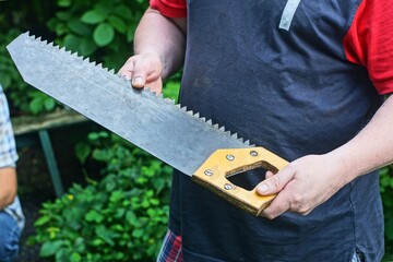 A man is holding a hand-held carpentry saw with a wooden handle outdoors. Close-up of a traditional hand tool used for sawing wood during construction, carpentry and repair work. © FEDOR