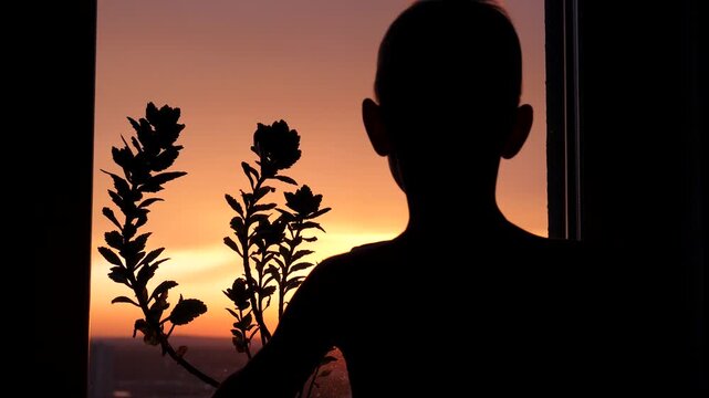 Silhouette of happy boy waking up at morning, stretching his arms and looking out window at beautiful orange sunrise. Kid enjoying moment of new day and hope at home. A child meets the dawn