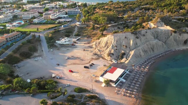 Cinematic aerial view looking down at the Kolymbia coast on Rhodes island, Greece. The footage captures luxury resort hotels, a coastal dry boat storage yard, rocky cliffs, a seaside tavern, and rows 
