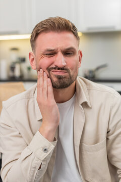 Young man at home gets sharp toothache covers cheek and grimaces then breathes slow for relief. Caucasian guy on sofa holds jaw with palm and shows pain worry and tense mood during hard wave and calm.