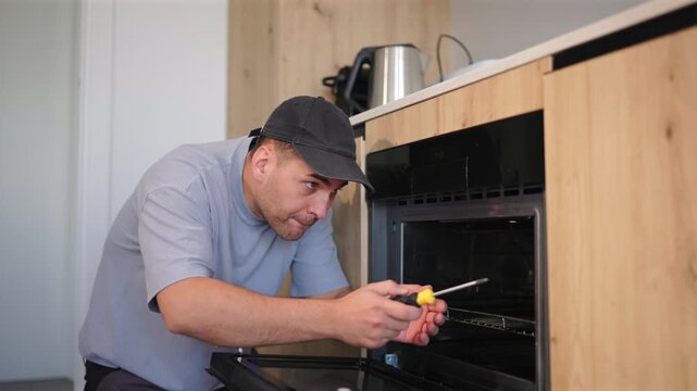 Professional repairman with toolbox performs maintenance and repair work on built-in oven in modern kitchen of home. Man wearing cap uses screwdriver to fix appliance door with focused concentration.