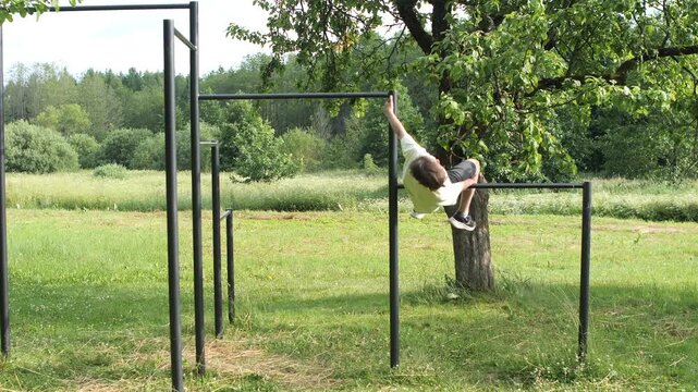 Young boy playing on outdoor fitness equipment bars and reaching for leaves in a lush green natural environment during a sunny summer day