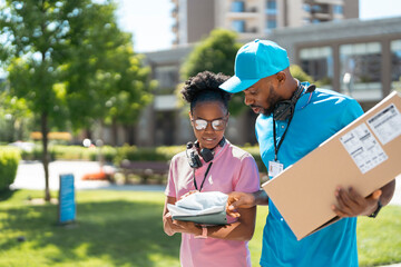 Two delivery workers meet outside in a green space. They check a package and a handheld device. The sun is shining and buildings are visible in the background. © Videophilia