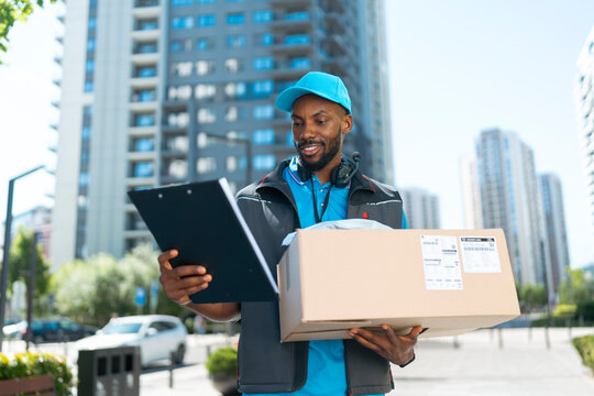 A delivery worker stands in a city street holding a package. He looks at a clipboard while wearing a blue cap and uniform. Tall buildings surround him in bright daylight.