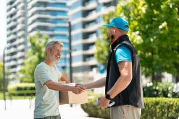 Naklejka premium A man receives a package from a delivery person in an urban setting. The two are outdoors near tall buildings and greenery on a sunny day. The smiling man shows appreciation.