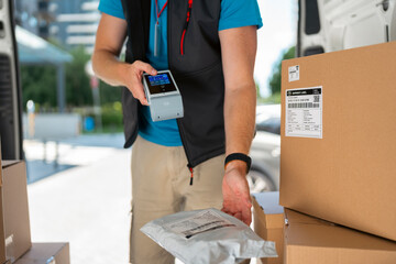 A delivery worker stands beside a van. He scans a package with a handheld device. Several boxes are around him. He is preparing to make a delivery on a busy street. © Videophilia