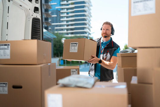 A delivery worker moves a large cardboard box from the back of a delivery van. He wears a headset and is surrounded by many other boxes in an urban setting.