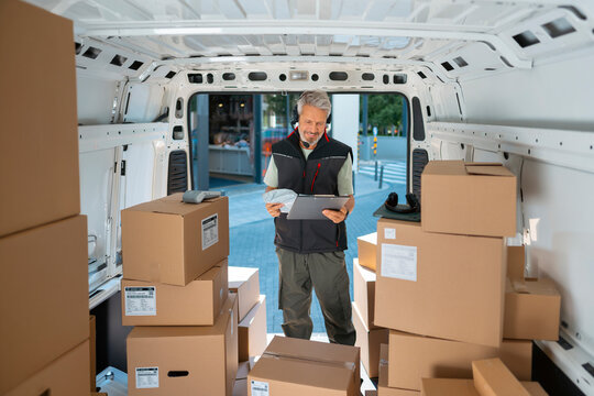 A delivery worker stands inside a van full of cardboard boxes. He holds a tablet and looks at the screen. The setting is during the day in a city area with some buildings in the background.