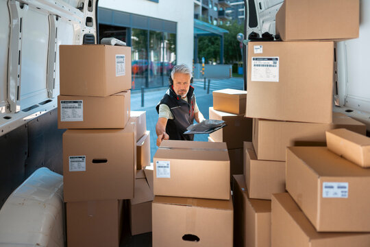 A delivery worker is inside a delivery van surrounded by many cardboard boxes. He is placing a box carefully in the space. It is a busy day for deliveries in the city.
