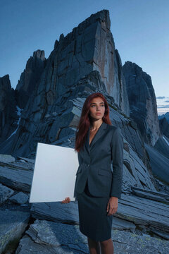 Woman with red hair stands against jagged mountain peaks holding a large white board under dim twilight sky, featuring sharp rock textures and cold blue tones in the composition.