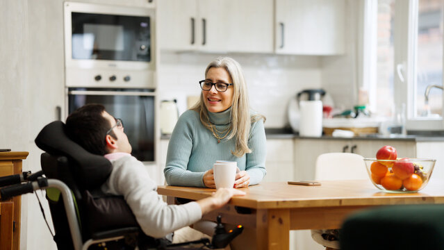 Woman and son talking, connecting, and bonding in a home kitchen. Man using a power chair, engaging with his mother