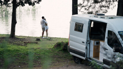 Couple hugging by a tranquil lake with their custom camper van parked nearby, embracing the freedom and romance of van life travel © DMegias