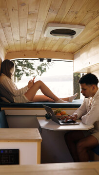 Young couple enjoying digital nomad lifestyle inside a custom camper van. Man working on laptop, woman relaxing with phone