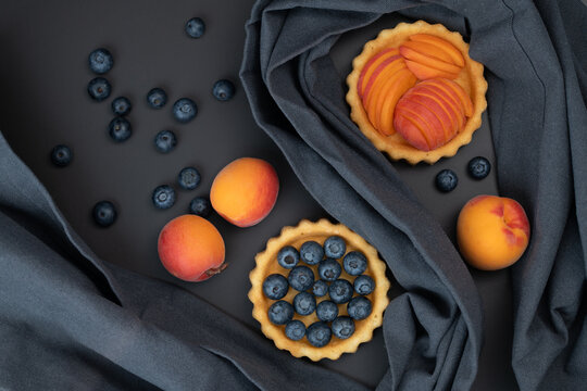 A top view of tartlets, which are small apricot cakes. One tartlet is topped with blueberries. Fruit is arranged around the cakes.