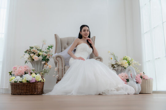 Wedding, Bride in beautiful dress sitting on sofa indoors in white studio interior, Young attractive Asian woman in wedding dress.