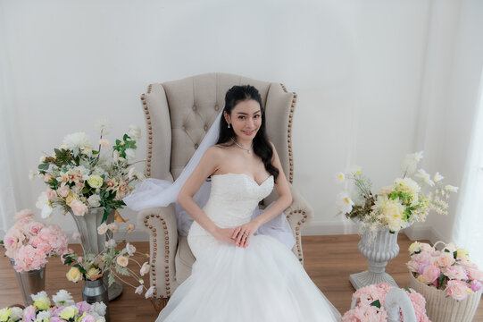 Wedding, Bride in beautiful dress sitting on sofa indoors in white studio interior, Young attractive Asian woman in wedding dress.