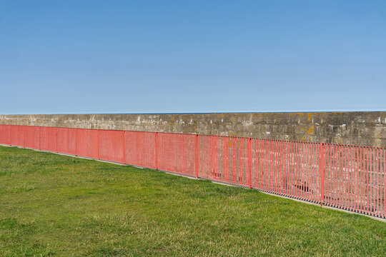A vibrant red metal fence stretches across green grass, bordered by a textured concrete sea wall under a bright blue sky.